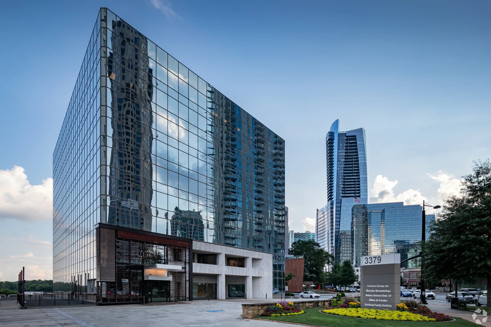 Modern glass office building reflecting city skyscrapers under a clear blue sky with manicured landscaping.